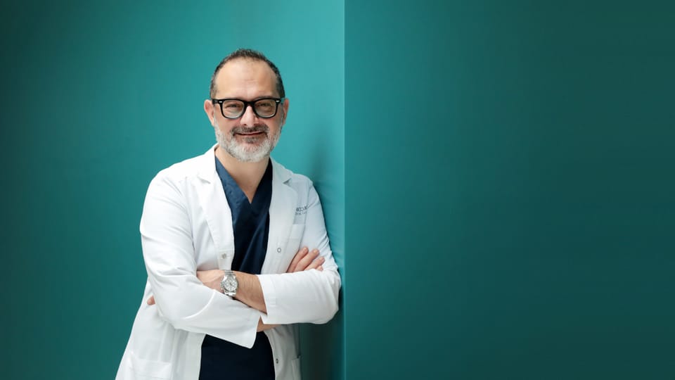 Il Prof. Luigi Coricciati in camice, a braccia conserte, sorride davanti a una parete color petrolio