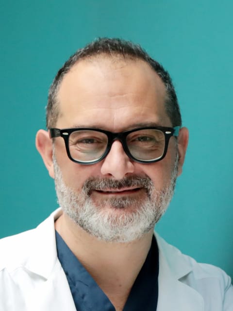 Il Prof. Luigi Coricciati in camice, a braccia conserte, sorride davanti a una parete color petrolio
